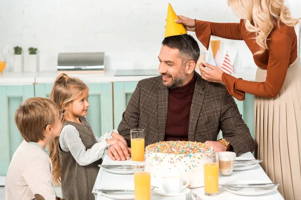 Frau setzt Partymütze auf glücklichen Mann, der neben Kindern am Küchentisch sitzt — Stockfoto