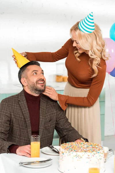 Smiling woman putting party cap on happy husband sitting at table with birthday cake — Stock Photo