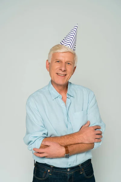 Happy senior man in party cap standing with crossed arms and smiling at camera isolated on grey — Stock Photo