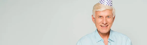 Panoramic shot of happy senior man in party cap smiling at camera isolated on grey — Stock Photo