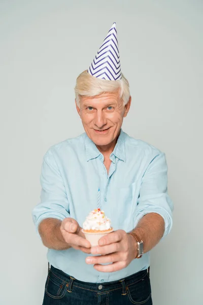 Happy senior man in party cap holding birthday cupcake and smiling at camera isolated on grey — Stock Photo