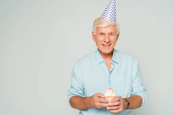 Happy senior man in party cap holding birthday cupcake and smiling at camera isolated on grey — Stock Photo