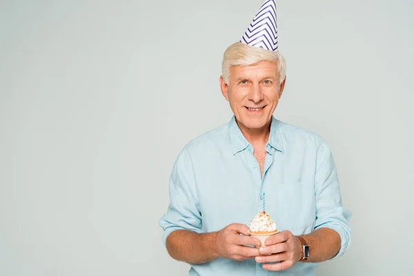 Feliz hombre mayor en partido gorra celebración cumpleaños cupcake y sonriendo a la cámara aislado en gris - foto de stock