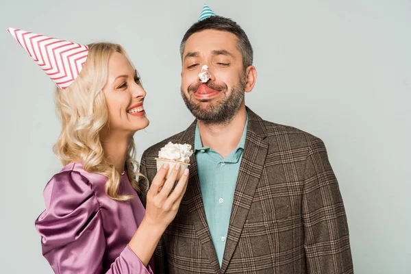Glückliche Frau hält Geburtstagskuchen in der Hand, während sie fröhlichen Mann mit Schlagsahne auf der Nase ansieht, isoliert auf grau — Stockfoto