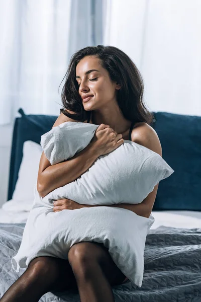 Улыбающаяся, мечтательная девушка, обнимающая подушку, сидя на кровати — стоковое фото