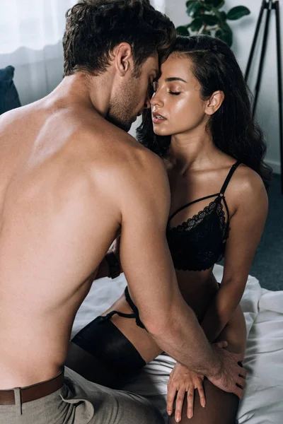 Homem sem camisa abraçando menina sedutora sentado na cama em lingerie preta — Fotografia de Stock