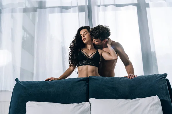 Sexy man kissing seductive girlfriend in black lingerie near window in bedroom — Stock Photo
