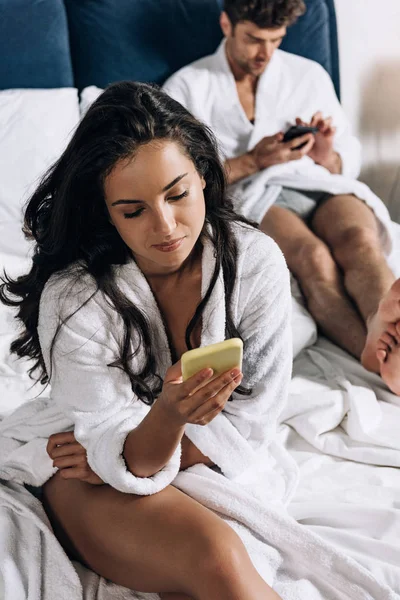 Selective focus of young couple in white bathrobes using smartphones in bedroom — Stock Photo