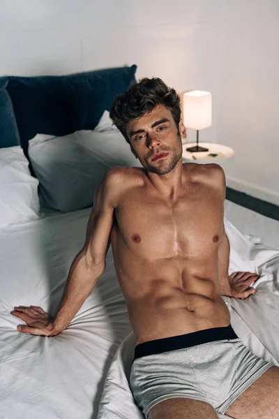 Sexy, pensoso uomo guardando fotocamera mentre seduto sul letto — Foto stock