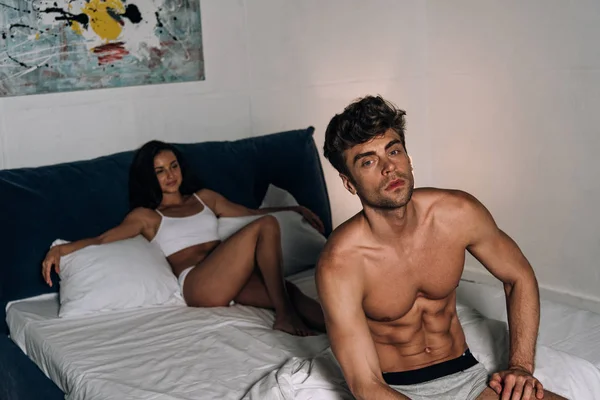 Pensive man looking at camera while sitting on bed near sexy girlfriend — Stock Photo