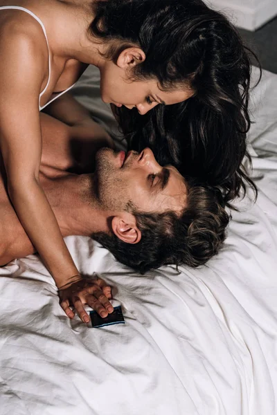 Sexy ragazza holding preservativo mentre sdraiato su uomo in letto — Foto stock