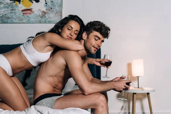 Feliz pareja en ropa interior sosteniendo vasos de vino tinto mientras está sentado en la cama - foto de stock