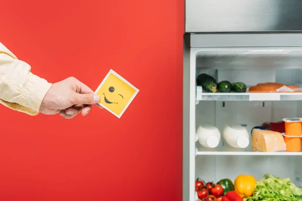 Vista cortada do homem segurando rosto sorridente perto da geladeira aberta com alimentos frescos em prateleiras isoladas em vermelho — Fotografia de Stock