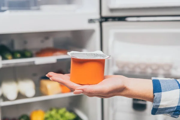 Vista cortada da mulher segurando iogurte perto da geladeira aberta com alimentos frescos nas prateleiras — Fotografia de Stock