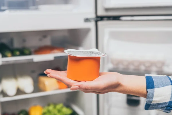 Vue recadrée de la femme tenant du yaourt près d'un réfrigérateur ouvert avec des aliments frais sur des étagères — Photo de stock