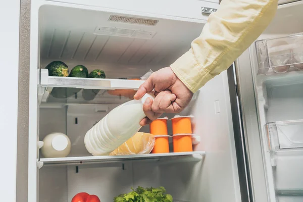 Vista recortada del hombre poniendo botella de leche en nevera con alimentos frescos en los estantes - foto de stock