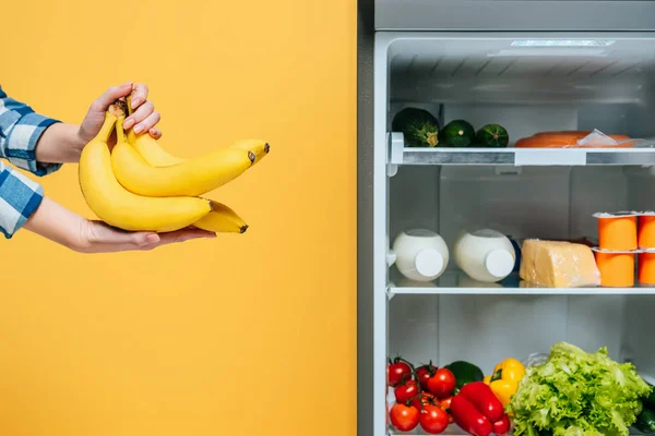 Vista recortada de la mujer sosteniendo plátanos cerca de nevera abierta con alimentos frescos en estantes aislados en amarillo - foto de stock
