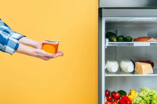 Обрезанный вид женщины, держащей йогурт рядом с открытым холодильником со свежими продуктами питания на полках изолированы на желтый — стоковое фото