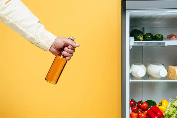 Abgeschnittene Ansicht eines Mannes, der eine Flasche Bier in der Nähe eines offenen Kühlschranks mit frischen Lebensmitteln in Regalen hält, isoliert auf gelb — Stockfoto