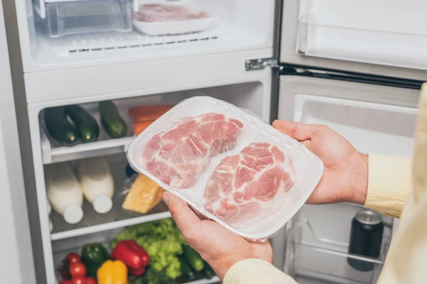 Vue recadrée de l'homme tenant de la viande congelée près d'un réfrigérateur ouvert plein de nourriture — Photo de stock