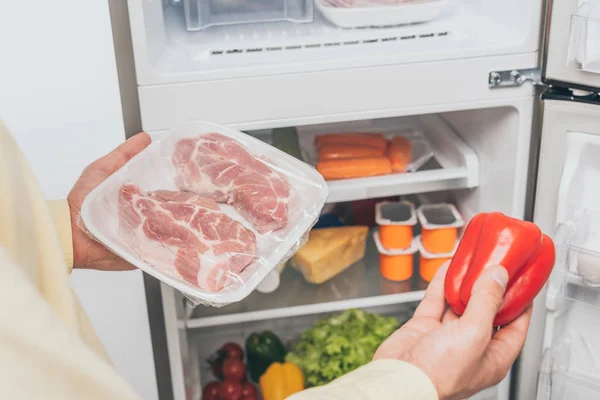 Cropped view of man holding frozen meat and fresh bell pepper near open fridge full of food — Stock Photo