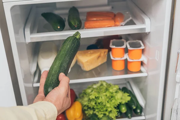 Cropped view of man holding cucumber near open fridge full of food — Stock Photo