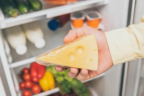 Vista recortada del hombre sosteniendo queso cerca de nevera abierta llena de alimentos - foto de stock