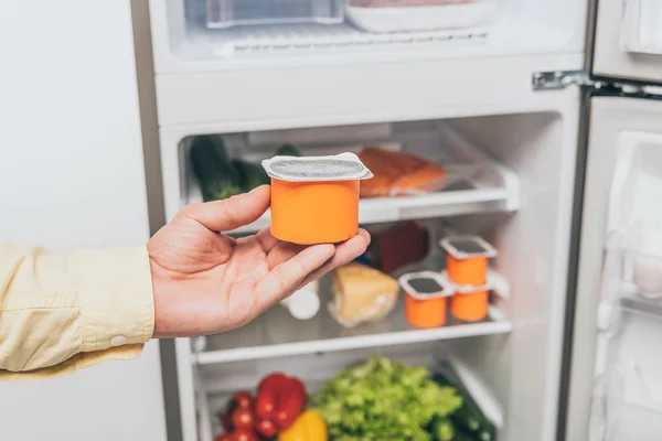 Vue recadrée de l'homme tenant du yaourt près d'un réfrigérateur ouvert plein de nourriture — Photo de stock