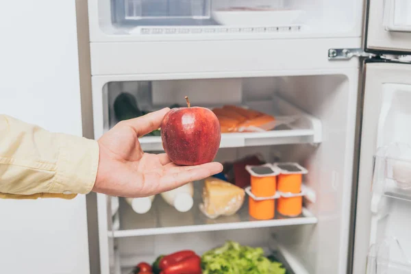 Cropped view of man holding red apple near open fridge full of food — Stock Photo