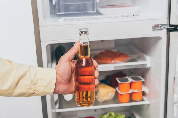 Vista cortada do homem segurando garrafa de cerveja perto da geladeira aberta cheia de alimentos — Fotografia de Stock