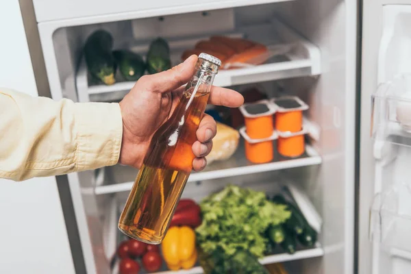 Cropped view of man holding bottle of beer near open fridge full of food — Stock Photo