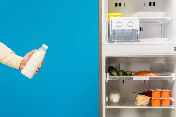 Обрізаний вид людини, що тримає молоко біля відкритого холодильника і морозильника зі свіжою їжею на полицях ізольовані на синьому — стокове фото