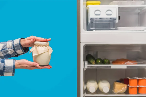 Vista recortada de la mujer sosteniendo crema agria cerca de nevera abierta y congelador con alimentos frescos en estantes aislados en azul - foto de stock