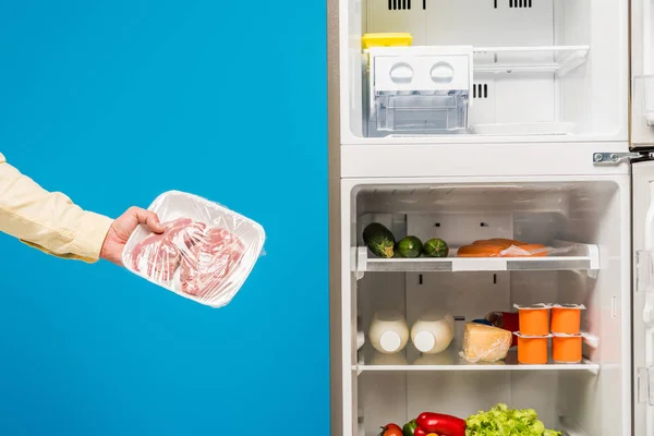Обрізаний вид людини, що тримає заморожене м'ясо біля відкритого холодильника та морозильника зі свіжою їжею на полицях ізольовані на синьому — стокове фото