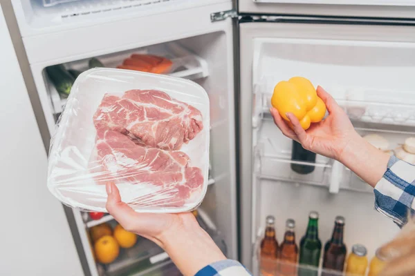 Vista cortada da mulher segurando carne congelada e pimentão perto da geladeira aberta com alimentos frescos nas prateleiras — Fotografia de Stock