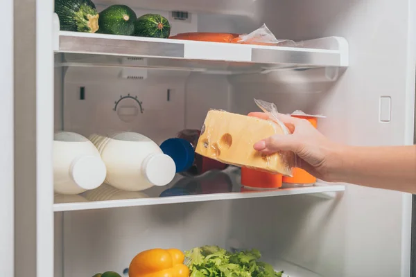 Vista cortada de mulher segurando queijo perto da geladeira aberta com alimentos frescos nas prateleiras — Fotografia de Stock
