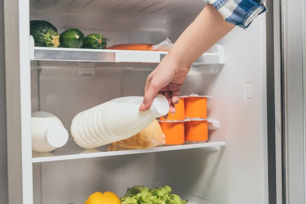 Cropped view of woman taking bottle of milk out from fridge with fresh food on shelves — Stock Photo