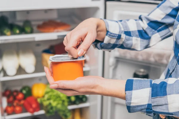 Cropped view of woman opening yogurt near open fridge with fresh food on shelves — Stock Photo