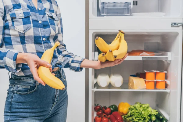 Vista cortada da mulher segurando bananas perto da geladeira aberta com alimentos frescos em prateleiras isoladas em branco — Fotografia de Stock