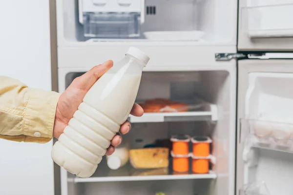 Vista ritagliata dell'uomo che tiene la bottiglia di latte vicino al frigorifero aperto con cibo fresco sugli scaffali isolati su bianco — Foto stock