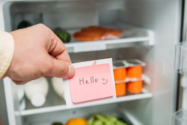 Vue recadrée de l'homme tenant une note collante avec lettrage bonjour près d'un réfrigérateur ouvert avec des aliments frais sur des étagères — Photo de stock