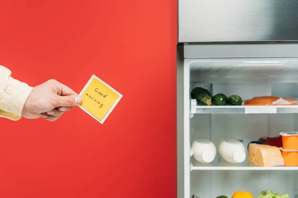 Vista ritagliata dell'uomo che tiene nota appiccicosa con scritte di buongiorno vicino al frigorifero aperto con cibo fresco sugli scaffali isolati sul rosso — Foto stock