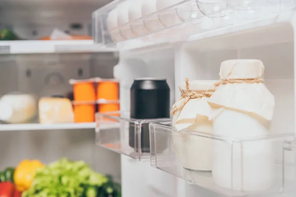 Focalizzazione selettiva di vasi con panna acida e lattina nera con soda sulla porta del frigorifero vicino al cibo fresco sugli scaffali — Foto stock