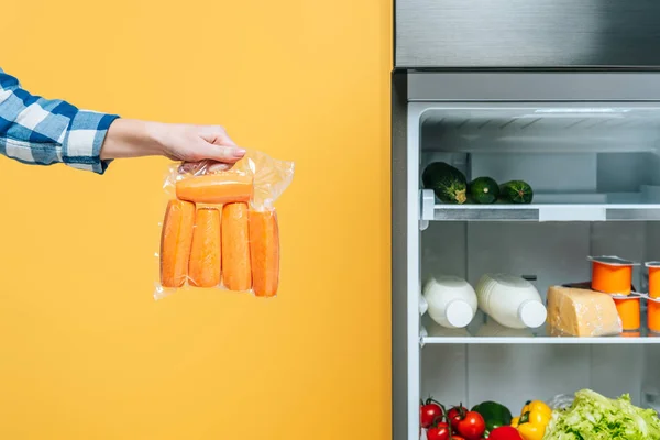 Vista ritagliata della donna che tiene le carote nel pacchetto vuoto vicino al frigorifero aperto con cibo fresco sugli scaffali isolati sul giallo — Foto stock