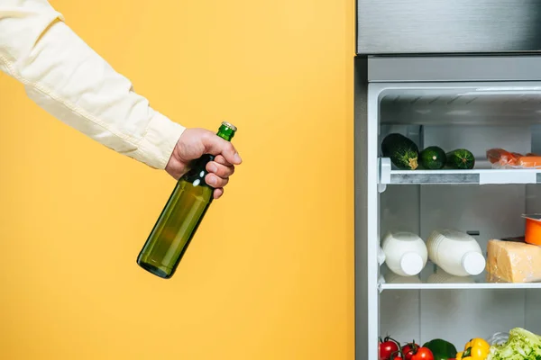 Vista recortada del hombre sosteniendo yogur cerca de nevera abierta con alimentos frescos en estantes aislados en amarillo - foto de stock