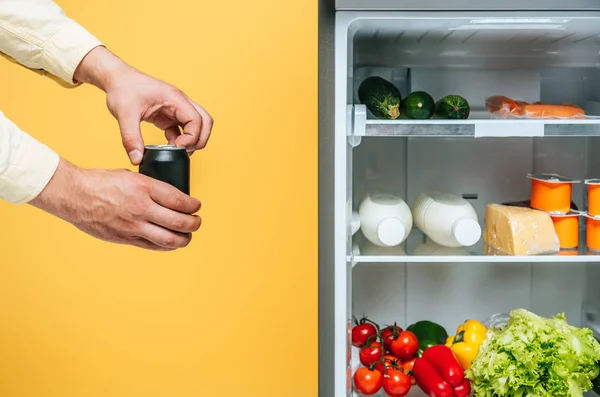 Abgeschnittene Ansicht eines Mannes, der eine Dose mit Limonade in der Nähe eines offenen Kühlschranks öffnet, mit frischen Lebensmitteln in den Regalen, die auf gelb isoliert sind — Stockfoto