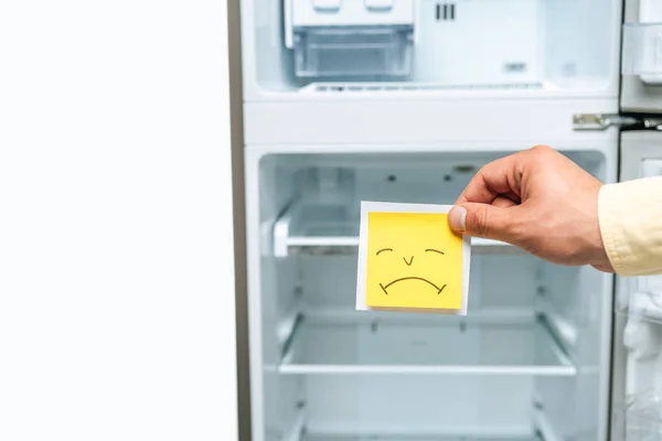 Recortado vista de hombre sosteniendo triste smiley cerca vacío abierto refrigerador aislado en blanco - foto de stock