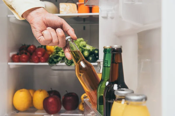 Vista cortada do homem tomando cerveja fora da geladeira com alimentos frescos nas prateleiras — Fotografia de Stock