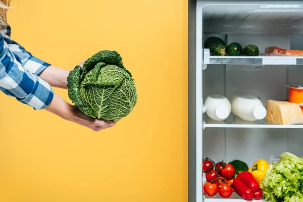 Abgeschnittene Ansicht einer Frau, die Kohl in der Nähe des offenen Kühlschranks mit frischen Lebensmitteln in Regalen hält, isoliert auf gelb — Stockfoto