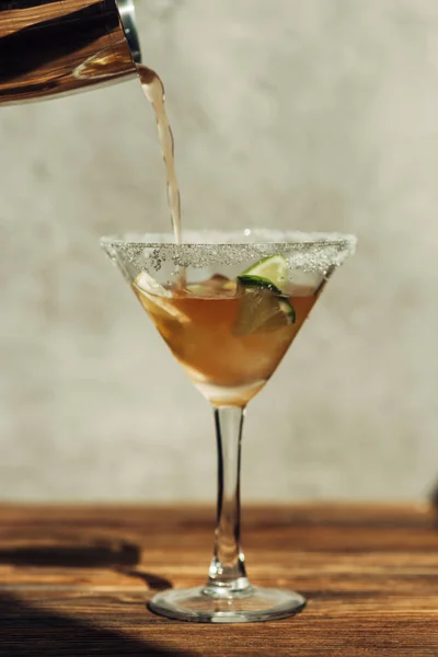 Liquid pouring from shaker into martini glass with ice, lemon and lime decorated with sugar on wooden surface in sunlight — Stock Photo